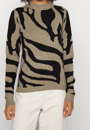 Frau trägt einen langärmligen Pullover mit beigem und schwarzem abstraktem Muster, kombiniert mit weißen Hosen, steht vor einem schlichten Hintergrund.