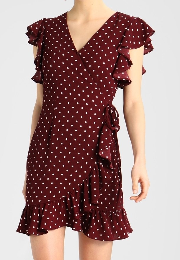 Robe portefeuille bordeaux à pois blancs, manches courtes à volants et ourlet à volants, nouée à la taille sur une femme debout.