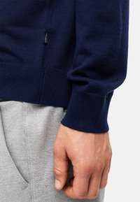 Maglione in maglia di colore blu navy con polsini e orlo a coste, realizzato in materiale morbido; pantaloni della tuta grigi parzialmente visibili sullo sfondo.