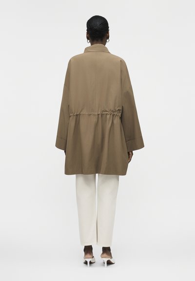 Žena s tmavou pokožkou a vlasmi zviazanými v drdole, oblečená v béžovom nadrozmernom kabáte so zúženým pásom, bielych nohaviciach a bielych topánkach na opätku, stojaca chrbtom.