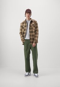 Brown plaid flannel shirt over a gray t-shirt, paired with green pants and black-and-white sneakers. Brown cap completes the look.