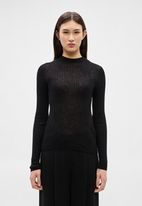 Sieviete ar taisniem tumšiem matiem nēsā piegulošu melnu ribotu garām piedurknēm topu un melnas bikses, stāvot pret baltu vienkāršu fonu.