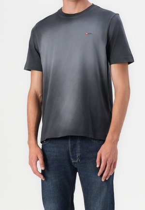 Mężczyzna ubrany w prostą ciemnoszarą koszulkę z krótkim rękawem z małym czerwonym logo na piersi oraz niebieskie dżinsy, stojący na białym tle.