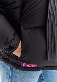 Czarna pikowana kurtka puchowa z fakturą przypominającą pikowania, z boczną kieszenią oraz czerwoną i niebieską metką. Założona na jasnoniebieskie dżinsy.