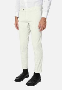 Pantaloni bianchi aderenti con una texture liscia, dotati di chiusura con un singolo bottone e passanti per la cintura, abbinati a scarpe nere e calzini.