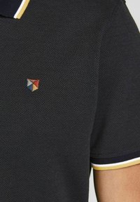 Navy polo shirt with textured fabric, yellow and white striped collar, featuring a small multicolored triangular logo on the chest.