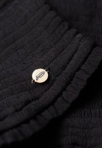 Nærbillede af sort ribbet stof med et lille rundt metalmærke indgraveret med ordet "Superdry" i kursiv skrifttype.