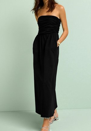Robe maxi noire sans bretelles avec un buste froncé, une taille plate, des poches latérales et un tissu lisse. Le modèle porte des sandales argentées à brides.