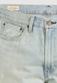 Jeans en denim bleu clair avec un design déchiré, style cinq poches, rivets en cuivre et une étiquette logo visible sur la ceinture.