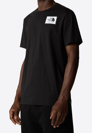 Mężczyzna w jednolitej czarnej koszulce z krótkim rękawem z białą prostokątną naszywką z logo North Face na lewej stronie klatki piersiowej.