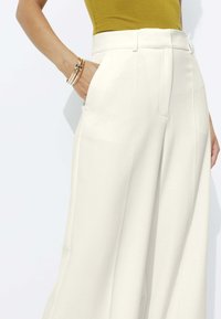 Pantalons blancs à jambes larges avec une texture lisse et des poches latérales, associés à un haut moutarde. Bras visible orné de bracelets fins.