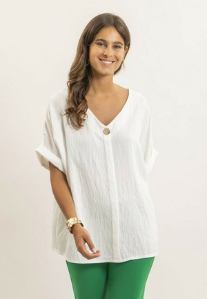 Femme souriante aux longs cheveux bruns portant un chemisier blanc ample aux manches retroussées et un pantalon vert, sur un fond clair.
