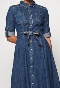 Denim button-up dress with chest pockets, rolled-up sleeves, and a tied belt at the waist, worn by a person standing against a neutral background.