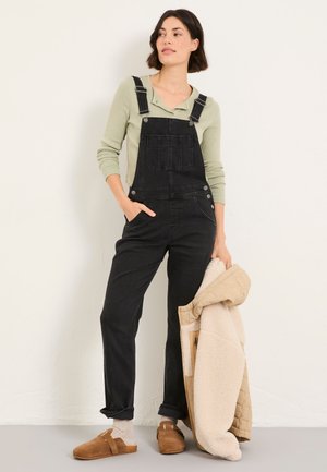 Frau trägt schwarze Jeanslatzhose über einem hellgrünen Langarmshirt, hält eine beigefarbene Lammfelljacke mit braunen Slippern und Socken.