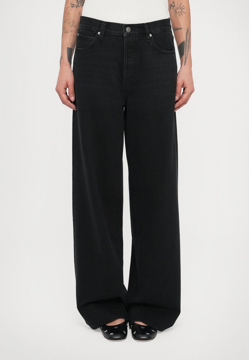 Személy magasderekú, bőszárú fekete farmert és fekete cipőt visel kivágásokkal, tetovált karok láthatók fehér háttér előtt.