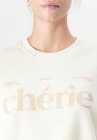 Primer plano de una persona que lleva un sudadera blanca con el texto beige "chérie" y frases más pequeñas "Art de Vivre" y "197 Rue du Magique París."
