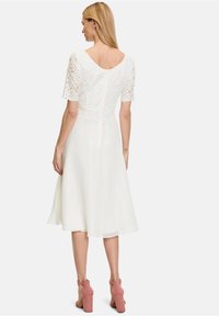 White dress featuring a lace bodice with short sleeves and a flared skirt. Back has a scoop neckline and a zipper. Worn with pink heeled sandals.