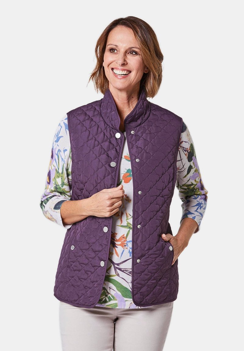 Lila gesteppte Weste mit hohem Kragen, ausgestattet mit Druckknöpfen und Seitentaschen, getragen über einem langärmligen Hemd mit floralem Muster.