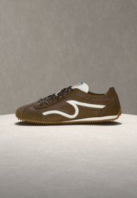 Χαμηλού προφίλ καφέ αθλητικό παπούτσι με μαύρα κορδόνια, λευκή κυματιστή πλευρική λωρίδα και υφή στην σόλα, σε ουδέτερη επιφάνεια και φόντο.