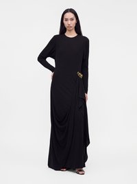 Woman with long dark hair wearing a full-length black dress with a gold brooch and open-toe black sandals against a plain white background.