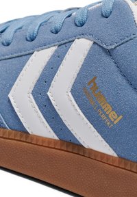 Baskets en daim bleu avec des bandes en chevron blanches, un logo doré et une semelle en caoutchouc marron texturée avec un motif en zigzag.