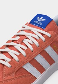 Κοραλί αθλητικό παπούτσι Adidas με λευκά κορδόνια, λευκές ρίγες στο πλάι, μπλε ετικέτα με λογότυπο στη γλώσσα και χρυσό κείμενο "NORA" στο πάνω μέρος.