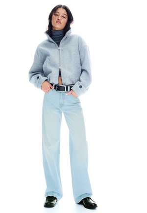 Nő szürke, rövid szabású cipzáros dzsekiben, sötét garbóban, világoskék, bőszárú farmerral, fekete övvel és fekete cipőben, zsebeiben tartja a kezét.