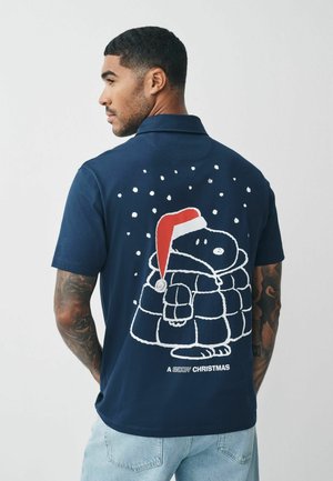 Jūras zila polo krekls ar baltu ilustrāciju, kas attēlo varoni ziemas ainavā ar sarkanu Ziemassvētku cepuri, sniegpārsliņām un tekstu "SNOOPY ZIEMASSVĒTKI".