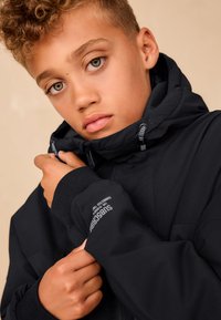 Musta takki, jossa on vetoketjullinen etuosa ja huppu, teksturoidulla kankaalla, joustavilla hihansuut ja logotunnus hihassa.