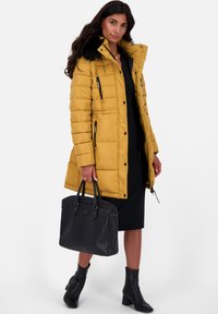 Vrouw in een gele gewatteerde jas met een zwarte neplederen kraag, zwarte jurk, zwarte enkellaarsjes, die een grote zwarte handtas vasthoudt.