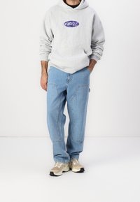 Szara bluza z kapturem z fioletowym logo graficznym, połączona z jasnoniebieskimi luźnymi dżinsami z detalami szwów oraz beżowymi sportowymi butami z czarnymi akcentami.