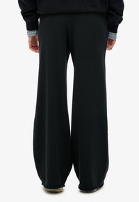 Persoon gekleed in losse zwarte broek, marineblauwe sweatshirt, en gestreepte mouwen zichtbaar, staande met ontspannen handen naast zich op een witte achtergrond.