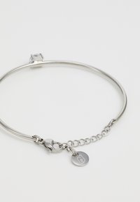 Silbernes Metallarmband mit glatter, runder Form, ausgestattet mit einem Kristallakzent und einer verbundenen Kette mit einem runden Logoetikett.