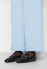 Schwarze Lederloafers mit goldfarbener Hardware-Akzent, die eine glatte Oberfläche und eine leicht abgerundete Spitze aufweisen, kombiniert mit hellblauen, weiten Hosen.