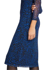 Žena oblečená v modro-černých vzorovaných šatech s přiléhavým střihem a poloprůhlednými rukávy, zobrazená od poloviny stehna po rameno, na bílém pozadí.