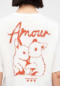 Δύο κόκκινα κουνέλια σε στυλ σκίτσου που κοιτάζονται μεταξύ τους, με τη λέξη "Amour" από πάνω και τη φράση "Connection Club" με τρεις καρδιές από κάτω σε ένα λευκό πουκάμισο.