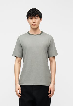 Jeune homme aux cheveux noirs courts portant un t-shirt gris à manches courtes et un pantalon noir, debout devant un fond blanc uni.