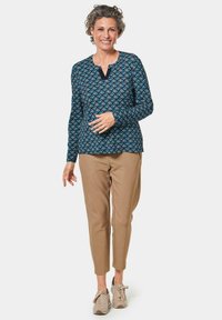 Langärmeliges blaues Oberteil mit einem Muster aus ineinandergreifenden Formen, kombiniert mit beigefarbenen, taillierten Hosen und hellen Sneakers.