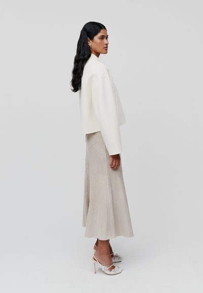 Witte cropped jacket, beige suède rok en transparante hoge hakken met verenaccenten, gepresenteerd tegen een neutrale achtergrond.