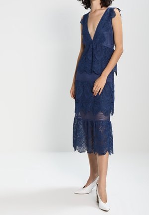 Granatowa koronkowa sukienka z głębokim dekoltem w kształcie litery V, falistymi krawędziami i wielowarstwowym dołem. Przezroczysta faktura i dopasowana sylwetka, zestawiona z białymi szpilkami.