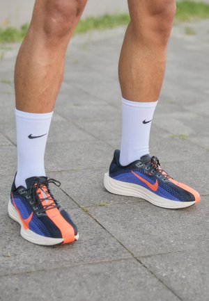 Piernas con calcetines blancos de Nike y zapatillas de correr Nike en negro, azul y naranja, paradas sobre un pavimento gris al aire libre.