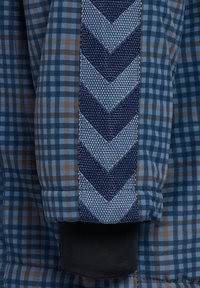 Blåt tjekmønsteret stof har en kontrasterende tekstureret navy-stribe med et chevron-design, afsluttet med en sort manchet i bunden.