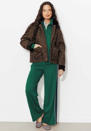 Femme portant une veste marron à imprimé léopard, un sweat à capuche vert zippé, un pantalon vert à jambes larges avec des bandes latérales, et des ballerines à imprimé léopard, debout contre un mur blanc.