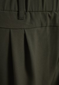 Tessuto verde scuro con una superficie liscia, caratterizzato da pieghe in vita e una cintura cucita. Il design è semplice e funzionale.