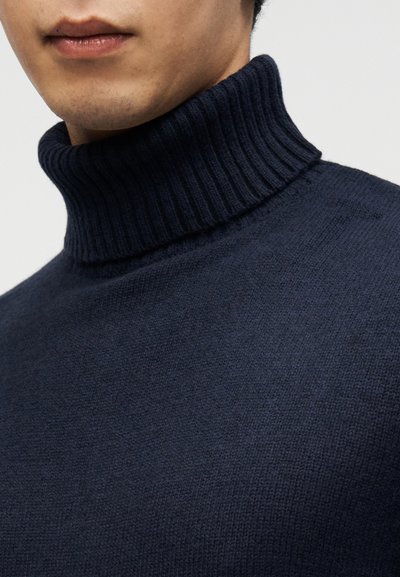 Σκούρο μπλε πουλόβερ με ψηλό ριμπ γιακά, απαλή υφή και εφαρμοστή γραμμή, που αναδεικνύει το λείο ύφασμα και το μινιμαλιστικό στυλ.