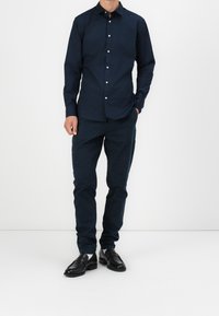 Camicia a maniche lunghe blu scuro con colletto a pulsante, abbinata a pantaloni scuri coordinati e scarpe in pelle nera. Tessuto morbido, vestibilità su misura.