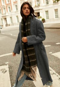 Manteau gris texturé avec des manches larges, associé à une écharpe sombre avec des rayures claires. Jeans bleu clair et arrière-plan d'une rue de la ville.