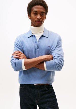 Giovane uomo con le braccia incrociate che indossa un maglione azzurro chiaro con colletto sopra un dolcevita bianco e jeans scuri, in piedi davanti a uno sfondo bianco semplice.