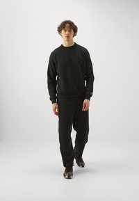 Sudadera negra y pantalones a juego en material suave con puños acanalados. Presenta detalles con cremallera en los laterales, combinados con zapatillas negras y grises.