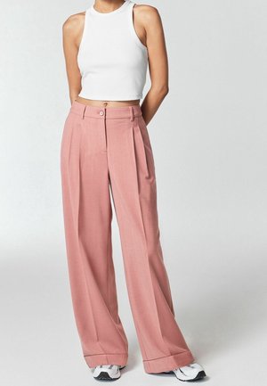 Frau trägt ärmelloses weißes Crop-Top, hochtaillierte, weit geschnittene rosa Hose und weiße Sneaker, steht mit den Händen hinter dem Rücken.